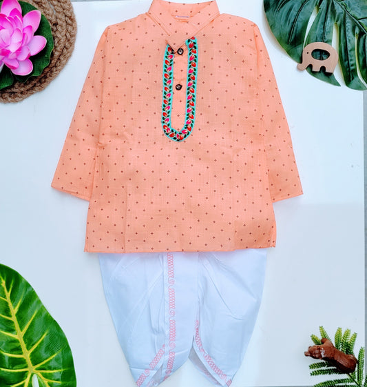 A kids' dhoti kurta set with a peach orange kurta and white dhoti. The kurta has small dark brown dots and a decorative green and blue necklace.